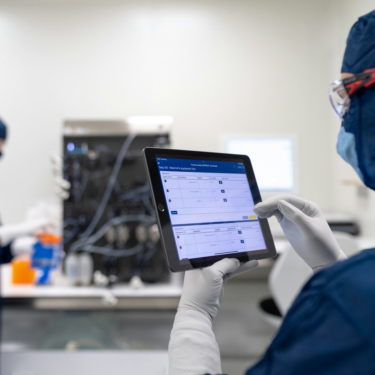 Teknisi berpakaian pelindung ruang bersih meninjau formulir terstruktur di tablet di dalam laboratorium, dengan rekan kerja yang bekerja di dekat peralatan di latar belakang