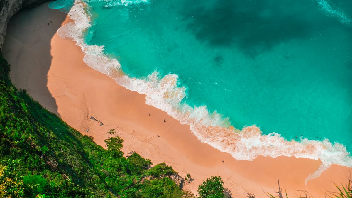 Aerial view of turquoise waves breaking on a sandy beach below green cliffs on the Bali coast